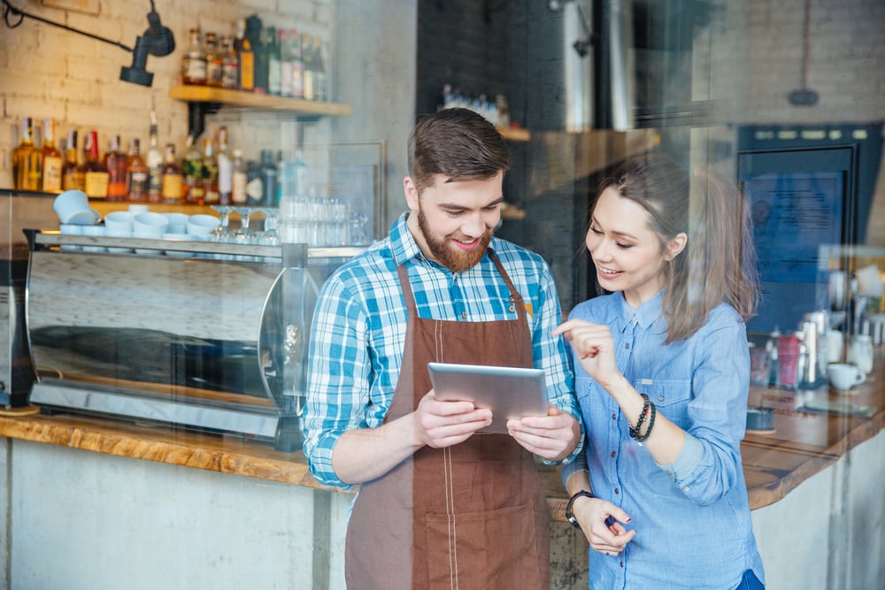 Un serveur souriant tient une tablette et une jeune femme la regarde dans un café.